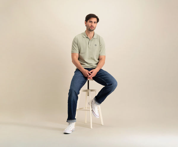 Een man met kort haar en een baard zit op een kruk tegen een neutrale achtergrond. Hij draagt "The N01 Polo Seagrass" en donkerblauwe jeans van Blue Rivet Jeans, gecombineerd met witte high-top sneakers, kijkt recht vooruit met een ontspannen houding.