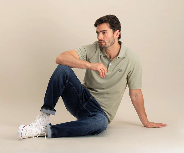 Een man die The N01 Polo Seagrass en Blue Rivet Jeans draagt, zit op de grond, één been gebogen en het andere gestrekt. Hij draagt witte high-top sneakers en kort haar en kijkt peinzend naar de zijkant tegen een effen beige achtergrond.