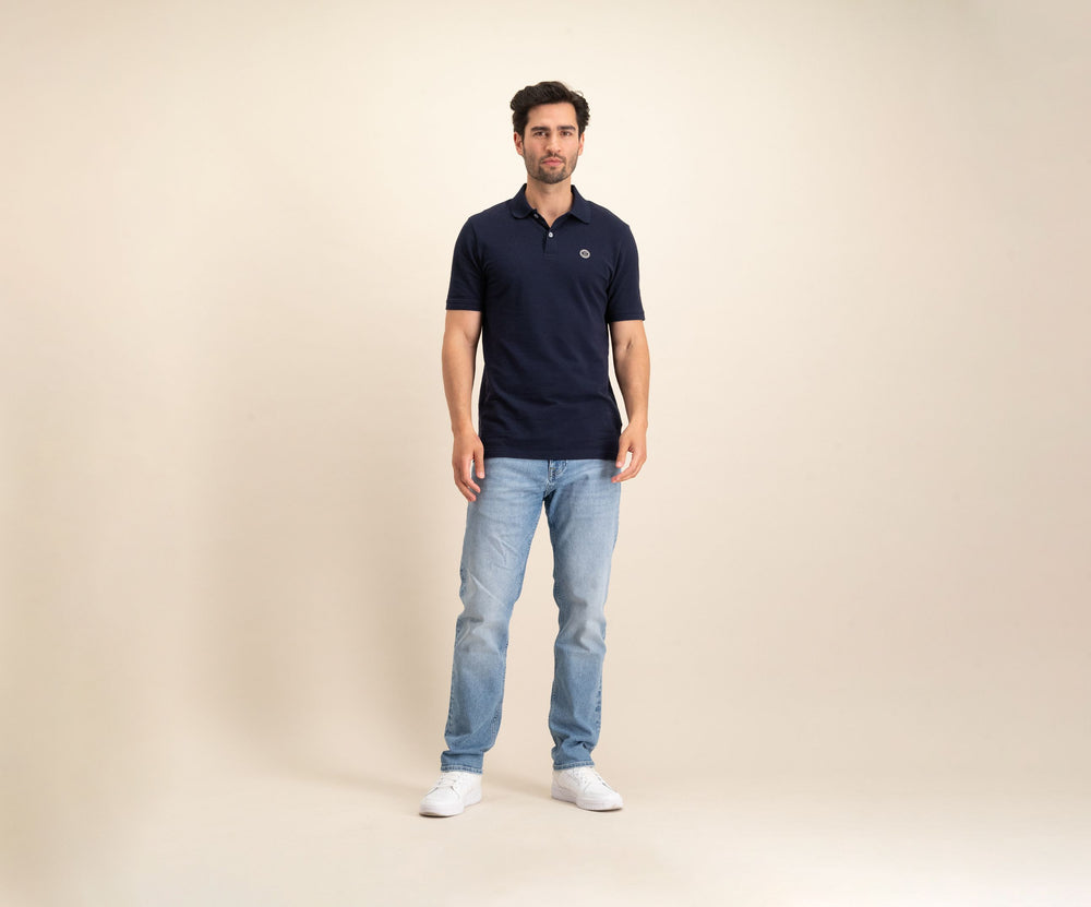 Tegen een effen beige achtergrond is een man te zien in The N01 Polo Navy en lichtblauwe Blue Rivet Jeans, aangevuld met witte sneakers. Hij heeft kort donker haar en een neutrale uitdrukking.