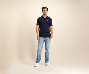 Tegen een effen beige achtergrond is een man te zien in The N01 Polo Navy en lichtblauwe Blue Rivet Jeans, aangevuld met witte sneakers. Hij heeft kort donker haar en een neutrale uitdrukking.