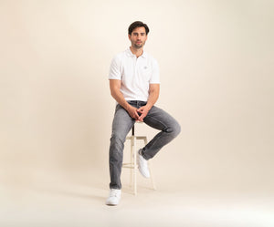 Een man die de N01 Polo White en grijze Blue Rivet Jeans draagt, samen met witte sneakers, zit op een kruk tegen een lichtbeige achtergrond. Hij lijkt ontspannen met één voet op de vloer en zijn handen rustend op zijn schoot.
