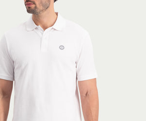 Een man draagt The N01 Polo White van Blue Rivet Jeans, met korte mouwen en een klein logo op de borst, staat tegen een lichte achtergrond. Zijn gezicht is gedeeltelijk zichtbaar en hij kijkt opzij.