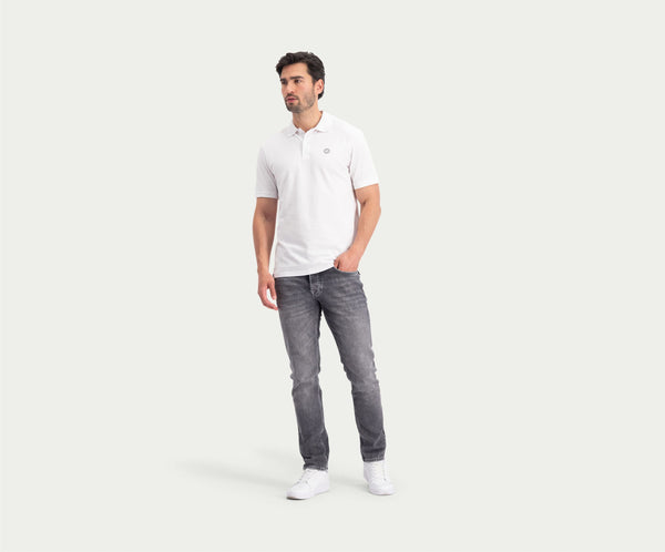 Een man staat in een ontspannen houding en draagt The N01 Polo White, gecombineerd met een grijze jeans van Blue Rivet Jeans. Met één hand in zijn zak maakt hij de look compleet met witte sneakers tegen een gebroken witte achtergrond.