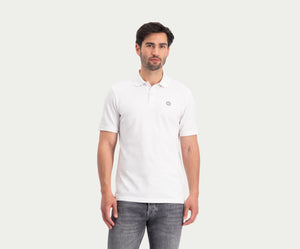 Een man die "The N01 Polo White" en Blue Rivet Jeans draagt, staat tegen een effen, lichte achtergrond. Hij heeft een neutrale uitdrukking met zijn armen langs zijn lichaam.