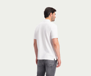 Een man draagt The N01 Polo White van Blue Rivet Jeans met korte mouwen en een grijze jeans en staat met zijn rug naar een lichte neutrale achtergrond.