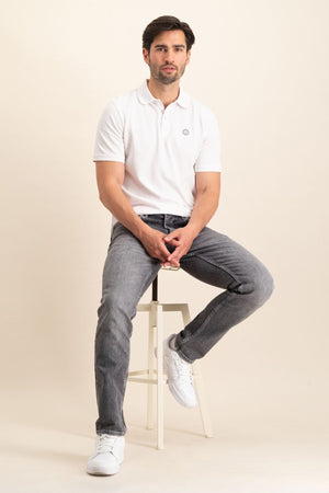Een man met donker haar zit op een kruk, draagt The N01 Polo White van Blue Rivet Jeans, grijze jeans en witte sneakers. De achtergrond is een effen, lichte kleur.