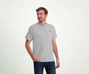 Een man met kort haar staat in de regular fit "The N01 Tee Grey Melee" en Blue Rivet Jeans. Hij heeft een baard en zijn rechterhand zit nonchalant in zijn zak tegen een effen witte muurachtergrond.