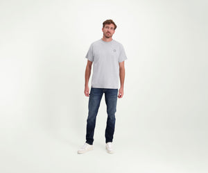 Een man die een The N01 Tee in Grey Melee draagt, staat tegen een effen achtergrond, gecombineerd met Blue Rivet Jeans en witte sneakers. Zijn korte, golvende haar complimenteert zijn ontspannen houding terwijl hij zelfverzekerd in de camera kijkt.
