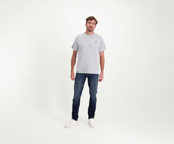 Een man die een The N01 Tee in Grey Melee draagt, staat tegen een effen achtergrond, gecombineerd met Blue Rivet Jeans en witte sneakers. Zijn korte, golvende haar complimenteert zijn ontspannen houding terwijl hij zelfverzekerd in de camera kijkt.