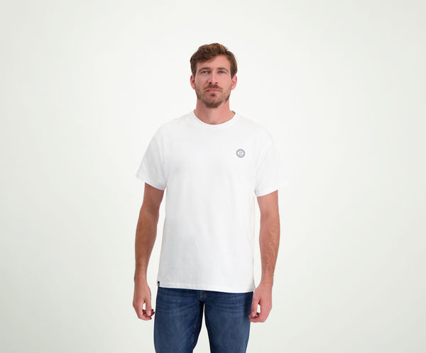 Tegen een effen lichte achtergrond straalt een man met kort bruin haar en een baard nonchalant zelfvertrouwen uit terwijl hij met zijn handen ontspannen langs zijn lichaam staat. Hij draagt The N01 Tee White van Blue Rivet Jeans, perfect gecombineerd met een blauwe jeans.