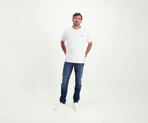 Een man in een regular fit The N01 Tee White and Blue Rivet Jeans staat tegen een effen witte achtergrond. Hij heeft zijn handen in zijn zakken en draagt witte sneakers.
