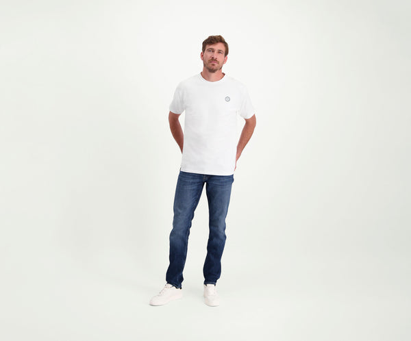 Een man in een regular fit The N01 Tee White and Blue Rivet Jeans staat tegen een effen witte achtergrond. Hij heeft zijn handen in zijn zakken en draagt witte sneakers.