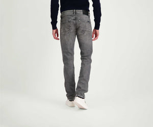 Een persoon wordt van achteren getoond met The N01 Slim Light Grey Used jeans van Blue Rivet Jeans, een donker shirt met lange mouwen en witte sneakers. Ze staan op een effen, lichtgekleurde achtergrond.