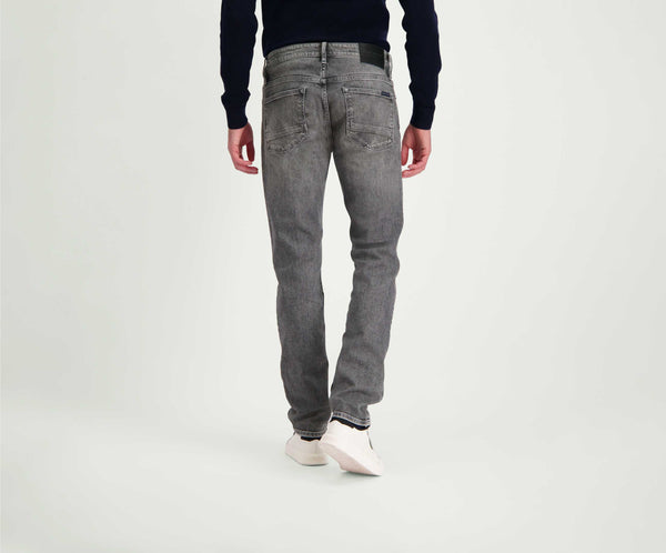Een persoon wordt van achteren getoond met The N01 Slim Light Grey Used jeans van Blue Rivet Jeans, een donker shirt met lange mouwen en witte sneakers. Ze staan op een effen, lichtgekleurde achtergrond.