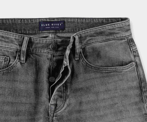 Close-up van de N01 Slim Light Grey Used denim jeans met een knoopsluiting, met een Blue Rivet Jeans-label op de tailleband. Deze slim jeans hebben een klassiek design met zakken aan de voorkant en een vervaagde wassing, en bieden precies de juiste hoeveelheid stretch voor comfort.