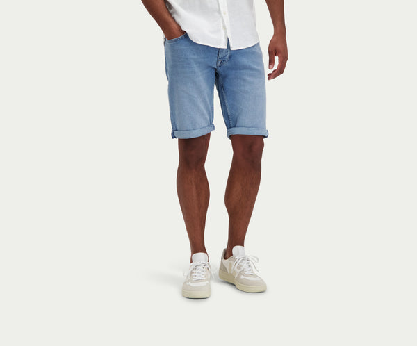 Een persoon die een wit shirt draagt, The N01 Short Stone Bleached by Blue Rivet Jeans, en witte sneakers staat nonchalant tegen een effen achtergrond. Hun handen zitten in de zakken van hun shorts, waarbij alleen de onderkant van hun lichaam zichtbaar is.