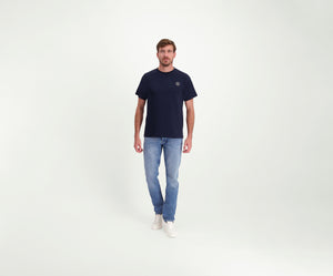 Een man draagt The N01 Tee Navy van Heren, lichtblauwe Blue Rivet jeans en regular-fit witte sneakers en staat tegen een effen witte achtergrond. Hij kijkt recht in de camera met een neutrale uitdrukking.
