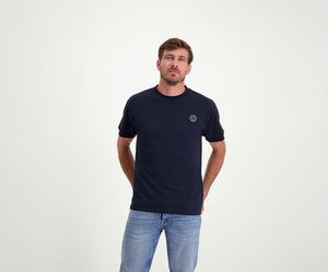 Een man met kort haar, gekleed in de kleuren The N01 Tee Navy en Blue Rivet Jeans, staat met zijn handen op zijn rug tegen een effen witte achtergrond.