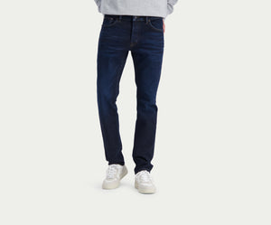 Een persoon die Blue Rivet Jeans' The N01 Slim Dark Indigo en witte sneakers draagt, staat tegen een effen lichte achtergrond. Het bovenlichaam is gedeeltelijk zichtbaar, met een vleugje lichtgrijs bovenwerk.