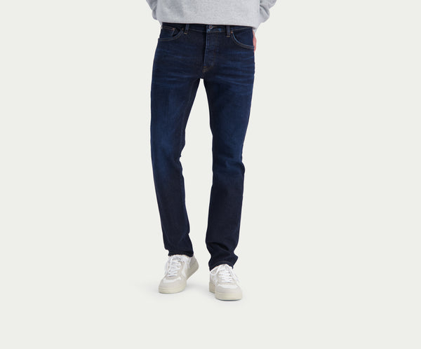 Een persoon die Blue Rivet Jeans' The N01 Slim Dark Indigo en witte sneakers draagt, staat tegen een effen lichte achtergrond. Het bovenlichaam is gedeeltelijk zichtbaar, met een vleugje lichtgrijs bovenwerk.