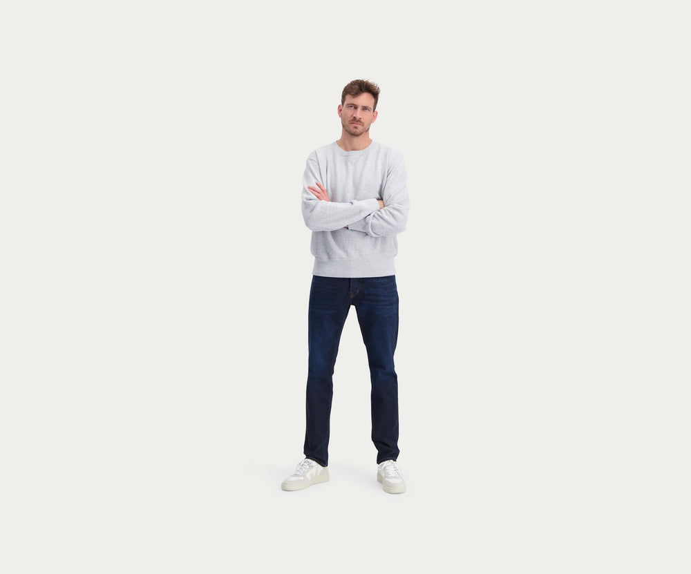 Een persoon die Blue Rivet Jeans' The N01 Slim Dark Indigo en witte sneakers draagt, staat tegen een effen lichte achtergrond. Het bovenlichaam is gedeeltelijk zichtbaar, met een vleugje lichtgrijs bovenwerk.