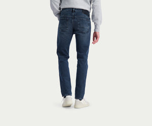 Een persoon die de N01 Slim Blue Black jeans van Blue Rivet Jeans draagt en een lichtgrijze sweater staat met zijn rug naar de camera. De premium jeans, gecombineerd met witte sneakers, bieden een relaxte look tegen de effen, lichtgekleurde achtergrond.