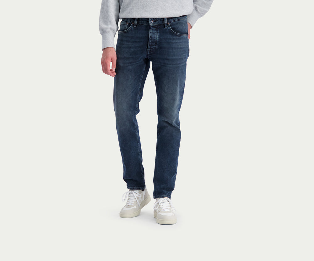 Een persoon die een paar The N01 Slim Blue Black jeans van Blue Rivet Jeans draagt, samen met een lichtgrijze sweater en witte sneakers, staat tegen een effen witte achtergrond. De afbeelding benadrukt de onderste helft van het individu, wat de strakke pasvorm van de jeans en strakke schoenen laat zien.