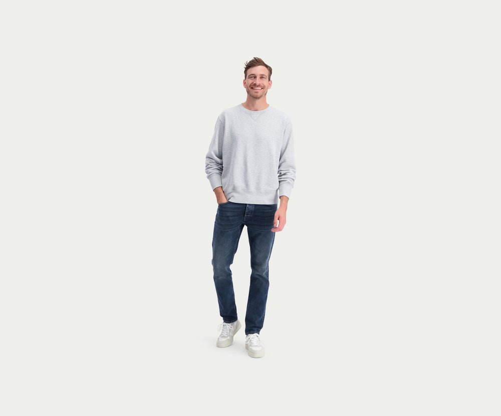 Een persoon die een paar The N01 Slim Blue Black jeans van Blue Rivet Jeans draagt, samen met een lichtgrijze sweater en witte sneakers, staat tegen een effen witte achtergrond. De afbeelding benadrukt de onderste helft van het individu, wat de strakke pasvorm van de jeans en strakke schoenen laat zien.
