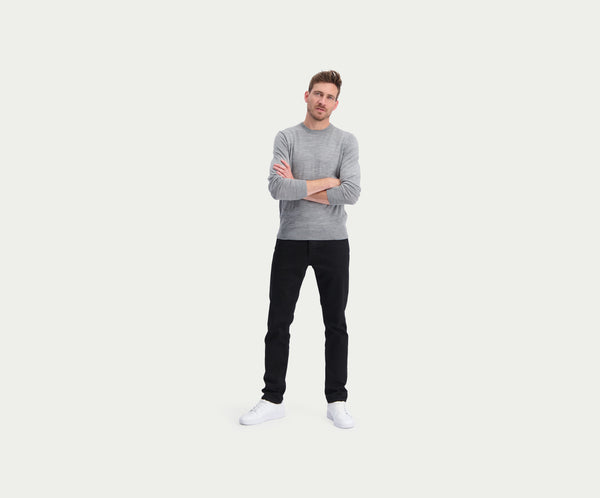 Een man staat met gekruiste armen, draagt een premium grijze trui, de N01 Slim Black broek van Blue Rivet Jeans en witte sneakers. Hij heeft kort haar en kijkt vooruit, tegen een effen witte achtergrond.