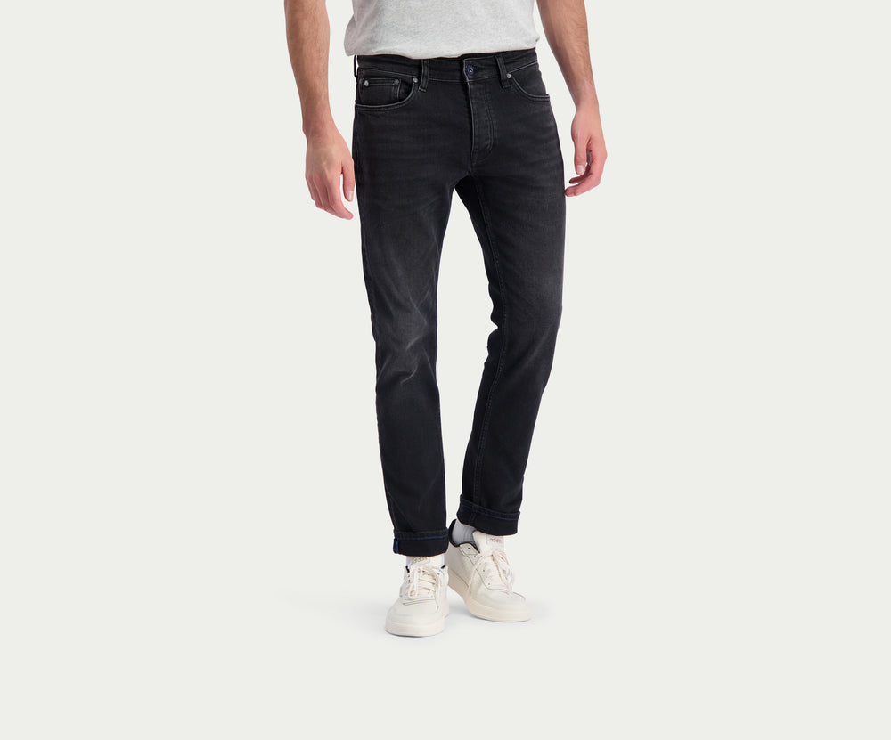 Een persoon die The N01 Slim Black Used jeans van Blue Rivet Jeans en witte sneakers draagt, staat tegen een effen lichte achtergrond. De zwarte jeans worden gecombineerd met een grijs t-shirt en de handen van de persoon liggen ontspannen langs hun zijden.