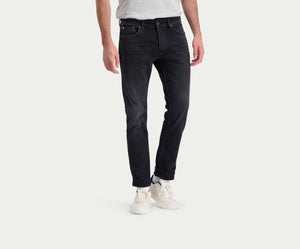Een persoon die The N01 Slim Black Used jeans van Blue Rivet Jeans en witte sneakers draagt, staat tegen een effen lichte achtergrond. De zwarte jeans worden gecombineerd met een grijs t-shirt en de handen van de persoon liggen ontspannen langs hun zijden.