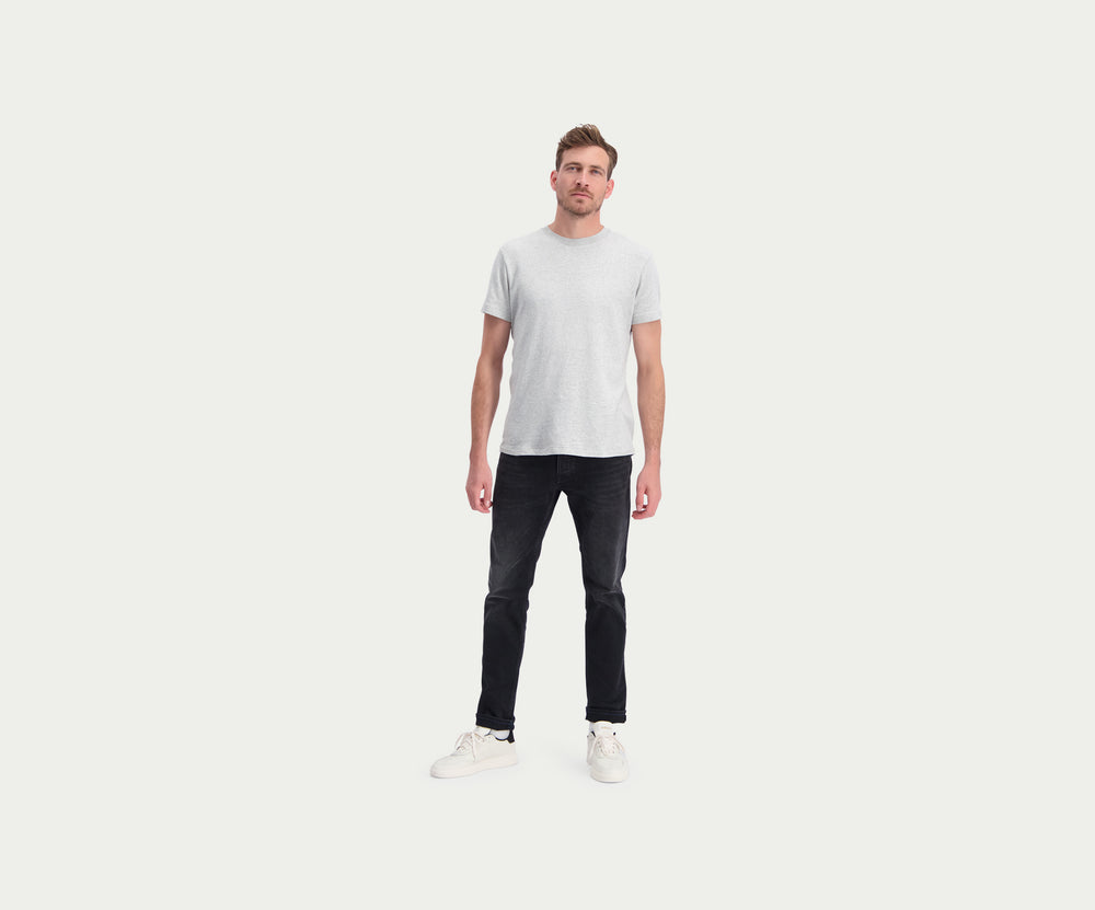Een persoon die The N01 Slim Black Used jeans van Blue Rivet Jeans en witte sneakers draagt, staat tegen een effen lichte achtergrond. De zwarte jeans worden gecombineerd met een grijs t-shirt en de handen van de persoon liggen ontspannen langs hun zijden.