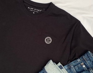 Een zwarte The N01 Tee van Blue Rivet Jeans, met een cirkelvormig logo op de borst, ligt naast een blauwe jeans op een wit oppervlak. Het Regular Fit t-shirtlabel straalt "simple. solid. style" uit.