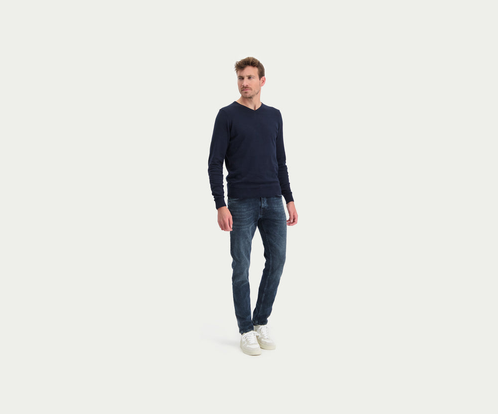 Een persoon gekleed in een donker shirt met lange mouwen en een The N01 Slim Blue Black Used jeans van Blue Rivet Jeans staat met zijn armen langs zijn lichaam en toont witte sneakers tegen een effen, lichtgekleurde achtergrond.