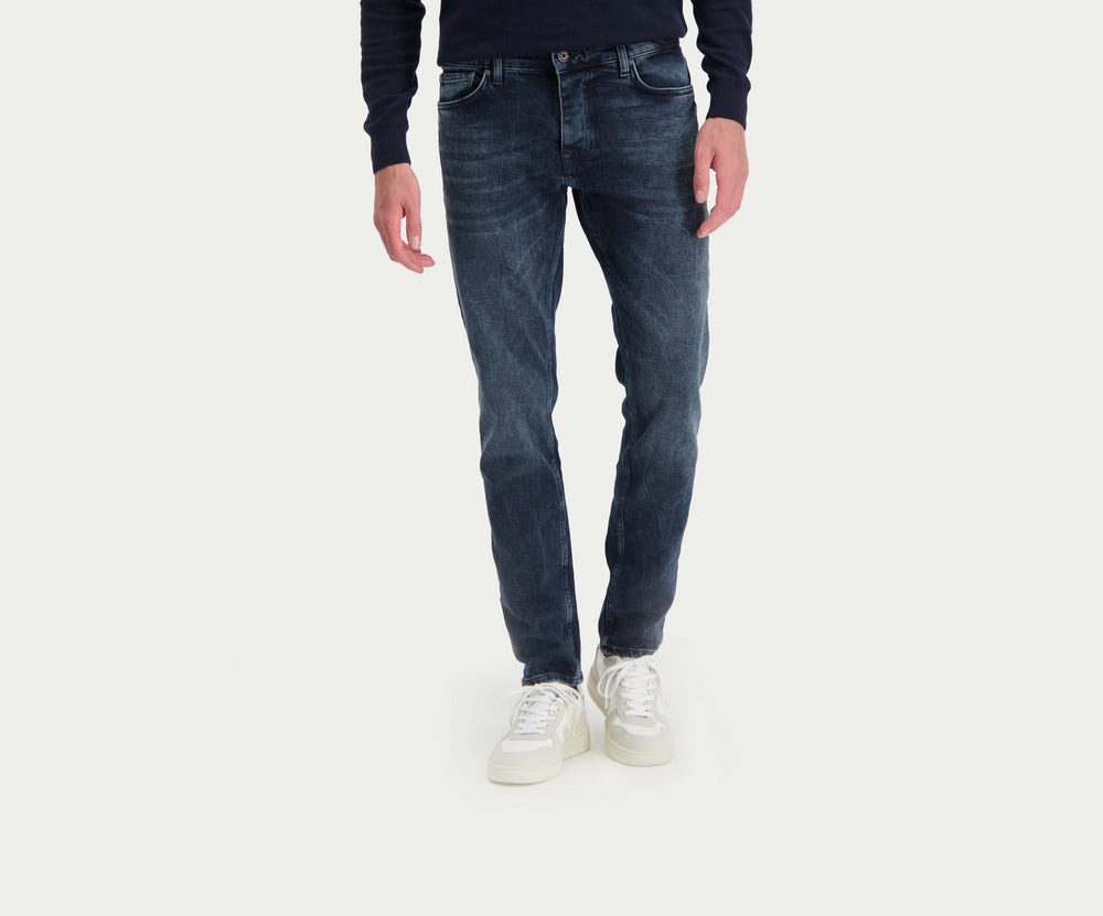 Een persoon gekleed in een donker shirt met lange mouwen en een The N01 Slim Blue Black Used jeans van Blue Rivet Jeans staat met zijn armen langs zijn lichaam en toont witte sneakers tegen een effen, lichtgekleurde achtergrond.