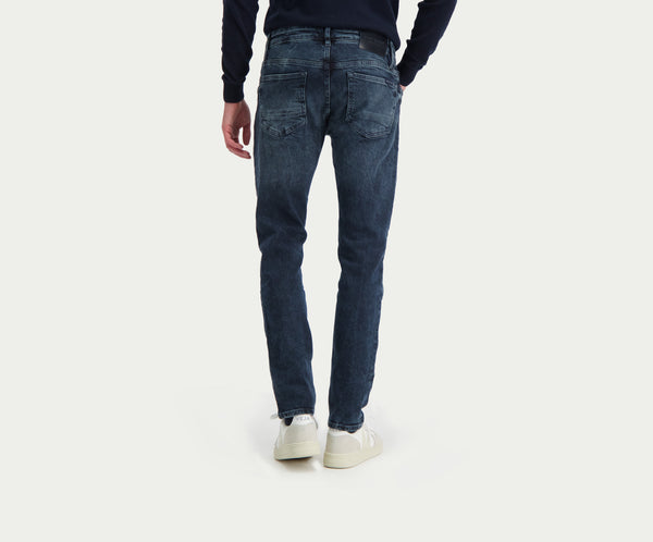 Een persoon draagt de N01 Slim Blue Black Used jeans van Blue Rivet Jeans en een donkere trui en staat met zijn rug naar de camera. Deze combinatie staat perfect bij witte sneakers tegen een lichtgekleurde achtergrond.
