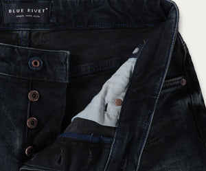 Close-up van donkerblauwe slim jeans met een zichtbare knoopsluiting. Het label luidt Blue Rivet Jeans met de tagline simple. solid. style. Koperen klinknagelknopen zijn prominent aanwezig en de binnenstof heeft een contrasterende lichtere kleur. De N01 Slim Blue Black Used spijkerbroeken bieden precies de juiste stretch voor comfort en pasvorm.