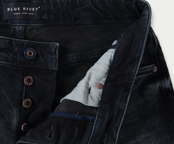 Close-up van donkerblauwe slim jeans met een zichtbare knoopsluiting. Het label luidt Blue Rivet Jeans met de tagline simple. solid. style. Koperen klinknagelknopen zijn prominent aanwezig en de binnenstof heeft een contrasterende lichtere kleur. De N01 Slim Blue Black Used spijkerbroeken bieden precies de juiste stretch voor comfort en pasvorm.