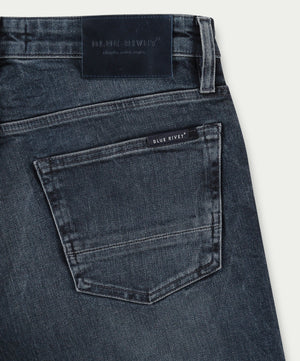 Een close-up van de N01 Slim Blue Black toont de eersteklas donkerblauwe denim jeans met een achterzak versierd met een klein label met de tekst "Blue Rivet Jeans". De tailleband is voorzien van een donker leren patch en de denim heeft een licht versleten textuur, wat de hoogwaardige vakmanschap benadrukt.
