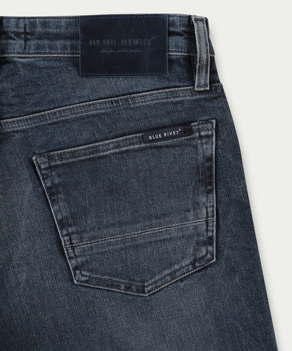 Een close-up van de N01 Slim Blue Black toont de eersteklas donkerblauwe denim jeans met een achterzak versierd met een klein label met de tekst "Blue Rivet Jeans". De tailleband is voorzien van een donker leren patch en de denim heeft een licht versleten textuur, wat de hoogwaardige vakmanschap benadrukt.