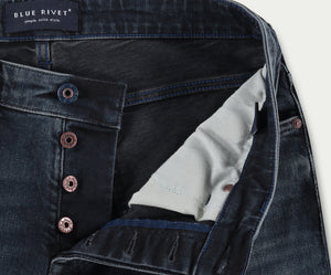 Close-up van premium donkere denim jeans met open gulp met knopen. Koperkleurige knopen en witte binnenvoering zijn zichtbaar. Het label luidt Blue Rivet Jeans met de tagline simple. solid. style. Perfect voor de spijkerbroekliefhebber die op zoek is naar kwaliteit en tijdloos design, dit zijn de N01 Slim Blue Black jeans.