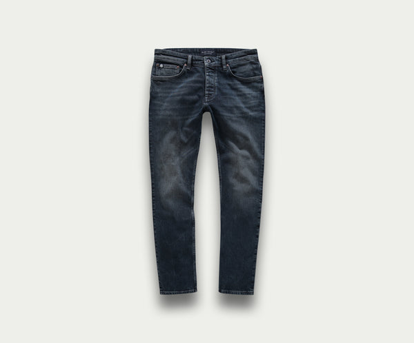 Introductie van de N01 Slim Blue Black van Blue Rivet Jeans: een premium paar slim-fit jeans in donkerblauw, met een subtiele fade op de dijen. Deze jeans zijn gemaakt met klassieke stiksels, vijf zakken en een knoopsluiting, elegant gepresenteerd tegen een lichtgrijze achtergrond.
