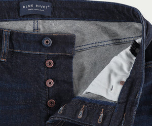 Close-up van de N01 Slim Dark Indigo jeans van Blue Rivet Jeans, met een knoopsluiting. De spijkerbroek is gedeeltelijk open om een lichtere contrasterende stoffen voering en een label aan de binnenkant van de tailleband te onthullen waarop Blue Rivet Jeans staat. Zichtbare stiksels omringen de knopen en zakken.