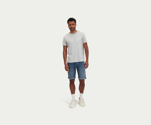 Een man staat tegen een effen witte achtergrond en draagt een lichtgrijs T-shirt, The N01 Short Dark Indigo Vintage van Blue Rivet Jeans, witte sokken en witte sneakers. Hij heeft krullend haar en een ontspannen houding.
