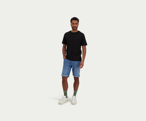 Een man staat tegen een effen achtergrond, draagt een zwart T-shirt gecombineerd met The N01 Short Indigo Used van Blue Rivet Jeans, groene sokken en witte sneakers. Zijn handen zitten nonchalant in zijn zakken.