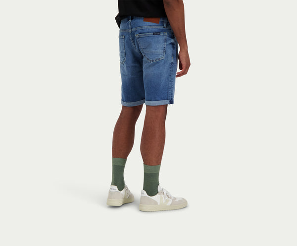 Een persoon die een zwart shirt draagt, The N01 Short Indigo Used by Blue Rivet Jeans, groene sokken en witte sneakers staat tegen een effen achtergrond. Het uitzicht is van achteren en iets naar de zijkant.