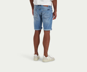 Een persoon staat in "The N01 Short Stone Bleached" denim shorts van Blue Rivet Jeans en witte sneakers, met het gezicht van de camera af. Ze dragen een wit T-shirt tegen een effen, lichtgekleurde achtergrond, waardoor hun stijlvolle shorts goed te zien zijn.