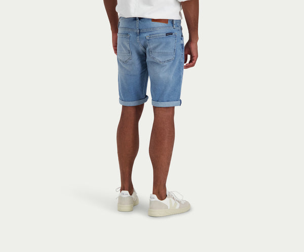 Een persoon staat in "The N01 Short Stone Bleached" denim shorts van Blue Rivet Jeans en witte sneakers, met het gezicht van de camera af. Ze dragen een wit T-shirt tegen een effen, lichtgekleurde achtergrond, waardoor hun stijlvolle shorts goed te zien zijn.