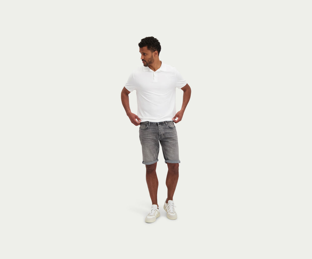 Een persoon die een wit t-shirt draagt, The N01 Short Light Grey Used by Blue Rivet Jeans, en witte sneakers staat tegen een effen achtergrond. De shorts zijn opgerold bij de zoom.