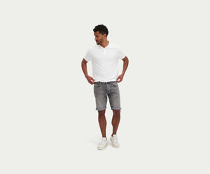 Een man met een wit poloshirt, The N01 Short Light Grey Used by Blue Rivet Jeans, en witte sneakers staat tegen een effen witte achtergrond. Hij kijkt naar voren met zijn handen op zijn heupen en kijkt lichtjes opzij.